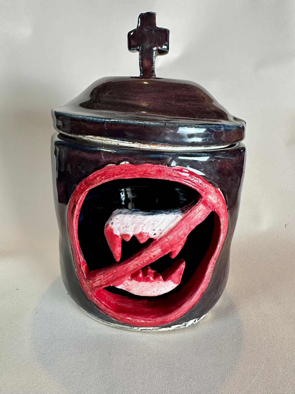 A glossy black canister with a cut out of a set of bloody vampire fangs inside a red circle with a slash; topped with a lid with a cross as a handle. Set against a solid neutral background.