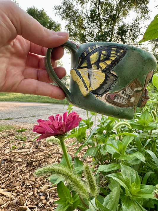 A hand holds a handmade green ceramic mug decorated with a yellow swallowtail butterfly and a partial brown Polyphemus moth; set against a garden background with a pink zinnia.