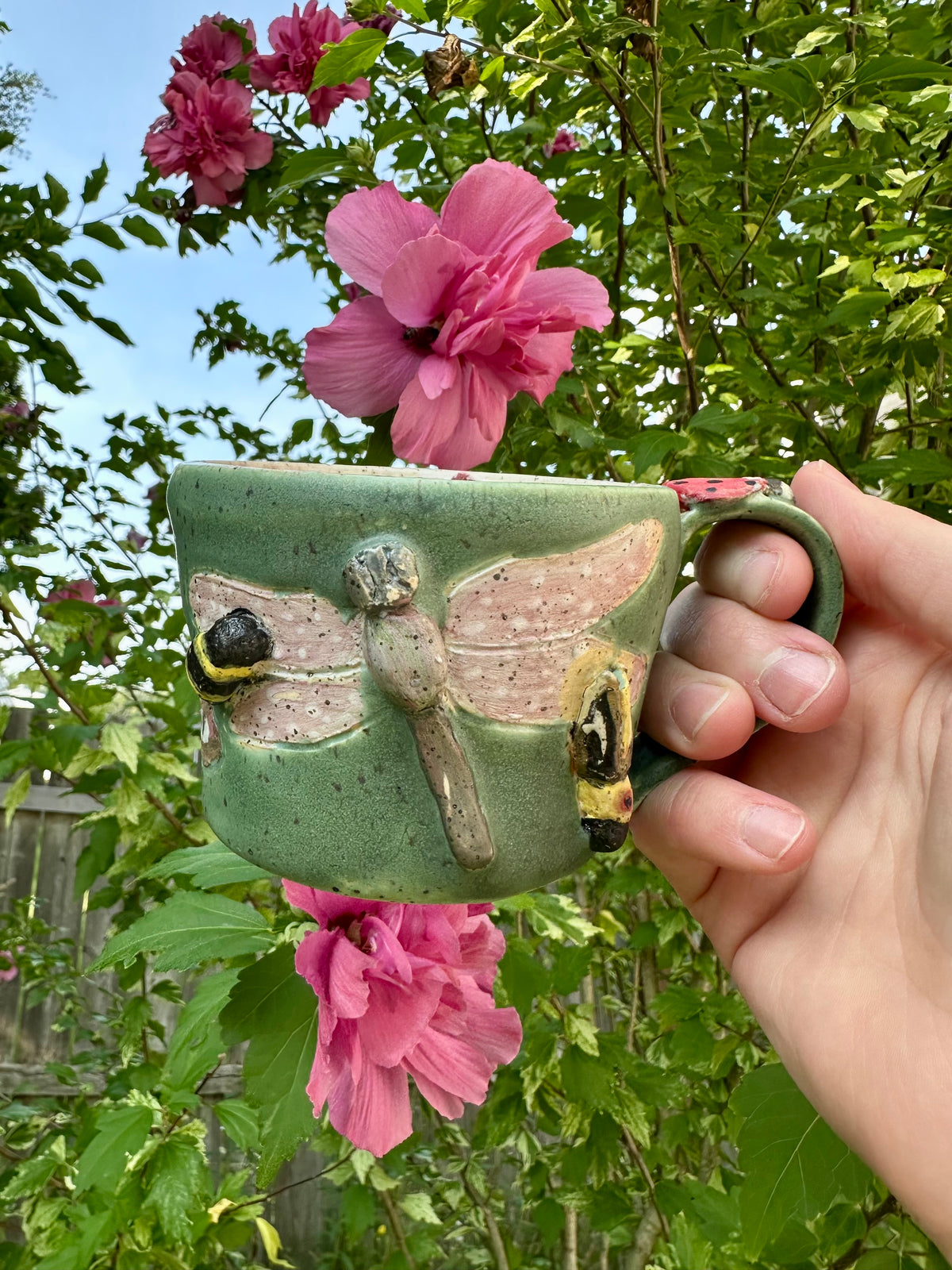 A handmade green ceramic mug decorated with a sculpted dragonfly, bumblebee, and firefly design is held by a hand by the handle; set against a background of green garden foliage and pink hibiscus flowers.