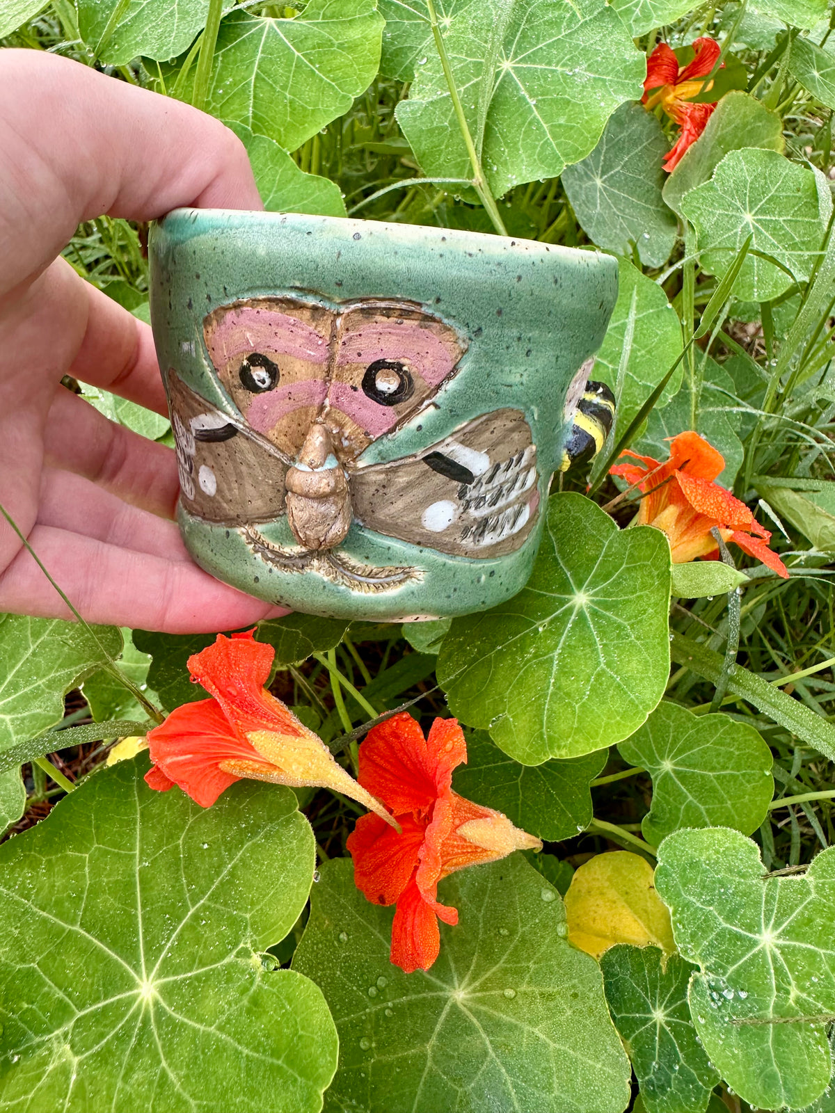 A hand holds a handmade green ceramic mug decorated with a brown Polyphemus moth and sculpted bumblebee; set against a garden background with orange nasturtiums.