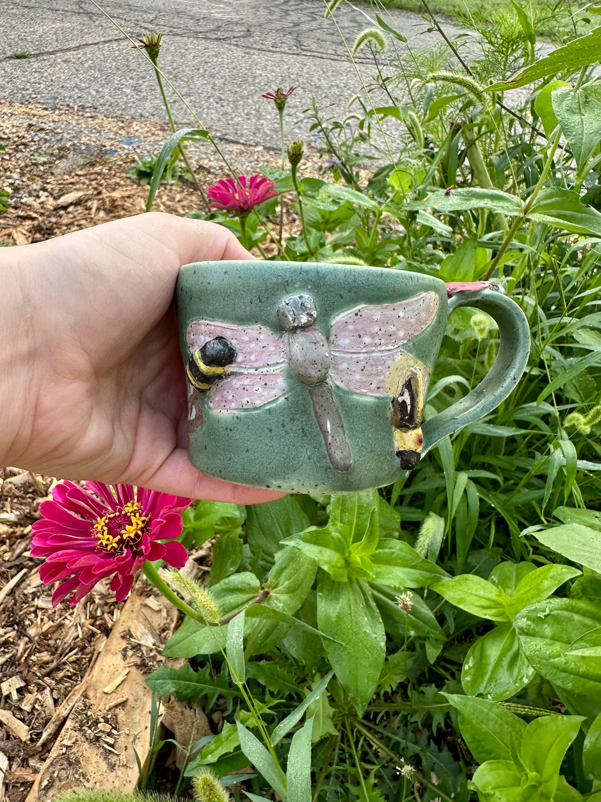 A hand holds a handmade green ceramic mug decorated with a bumblebee, purple and green dragonfly, and a firefly; set against a garden background with a pink zinnia.
