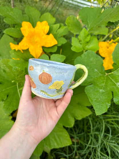 A hand holds a blue ceramic mug with illustrations of pumpkins, pumpkin flowers, and a green handle; set against a garden background of pumpkin leaves and pumpkin flowers.