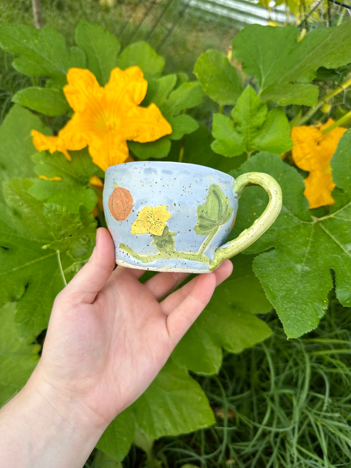 A hand holds a blue ceramic mug with illustrations of pumpkins, pumpkin flowers, pumpkin leaves, and a green handle; set against a garden background of pumpkin leaves and pumpkin flowers.