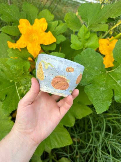 A hand holds a blue ceramic mug with illustrations of different kinds of pumpkins (small, large, warty, classic); set against a garden background of pumpkin leaves and pumpkin flowers.