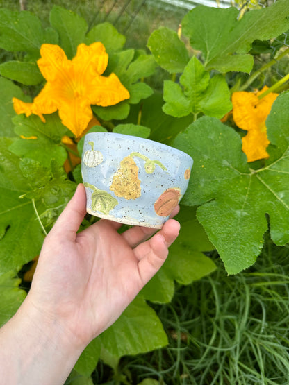 A hand holds a blue ceramic mug with illustrations of different kinds of pumpkins (large, warty, white) and pumpkin leaves; set against a garden background of pumpkin leaves and pumpkin flowers.