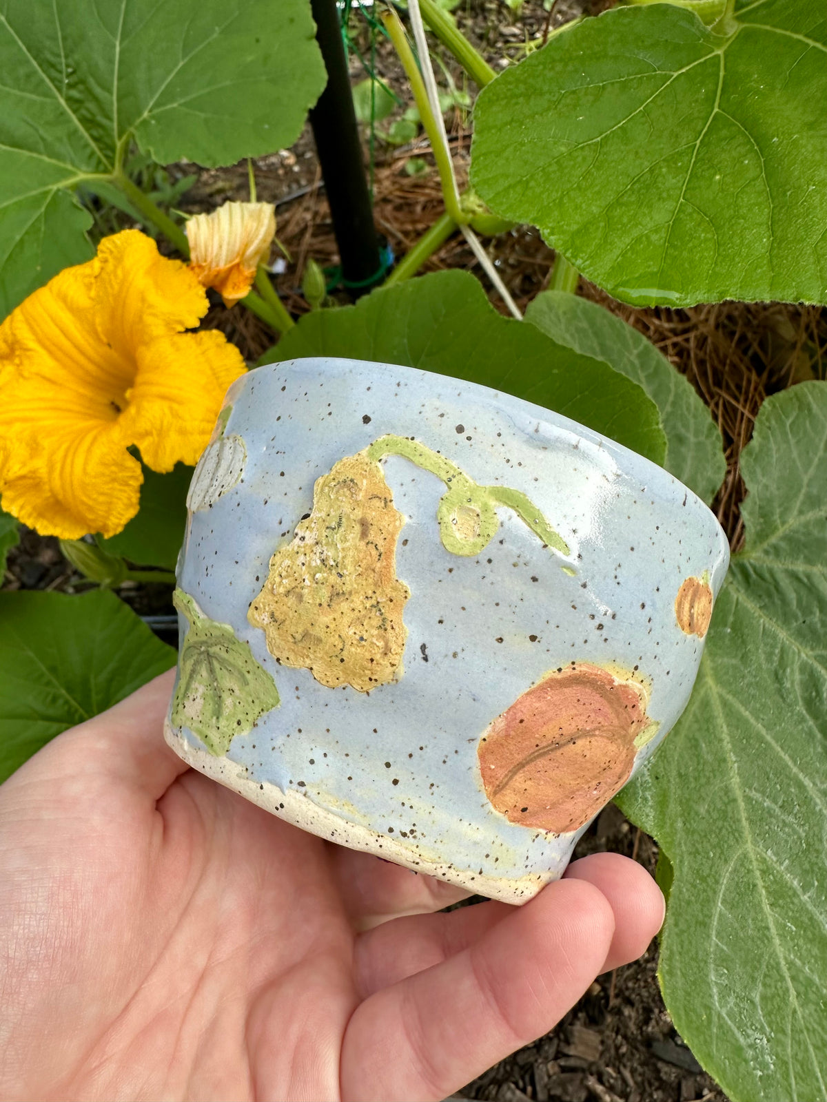 A hand holds a blue ceramic mug with illustrations of different kinds of pumpkins (large, warty, white) and pumpkin leaves; set against a garden background of pumpkin leaves and pumpkin flowers.