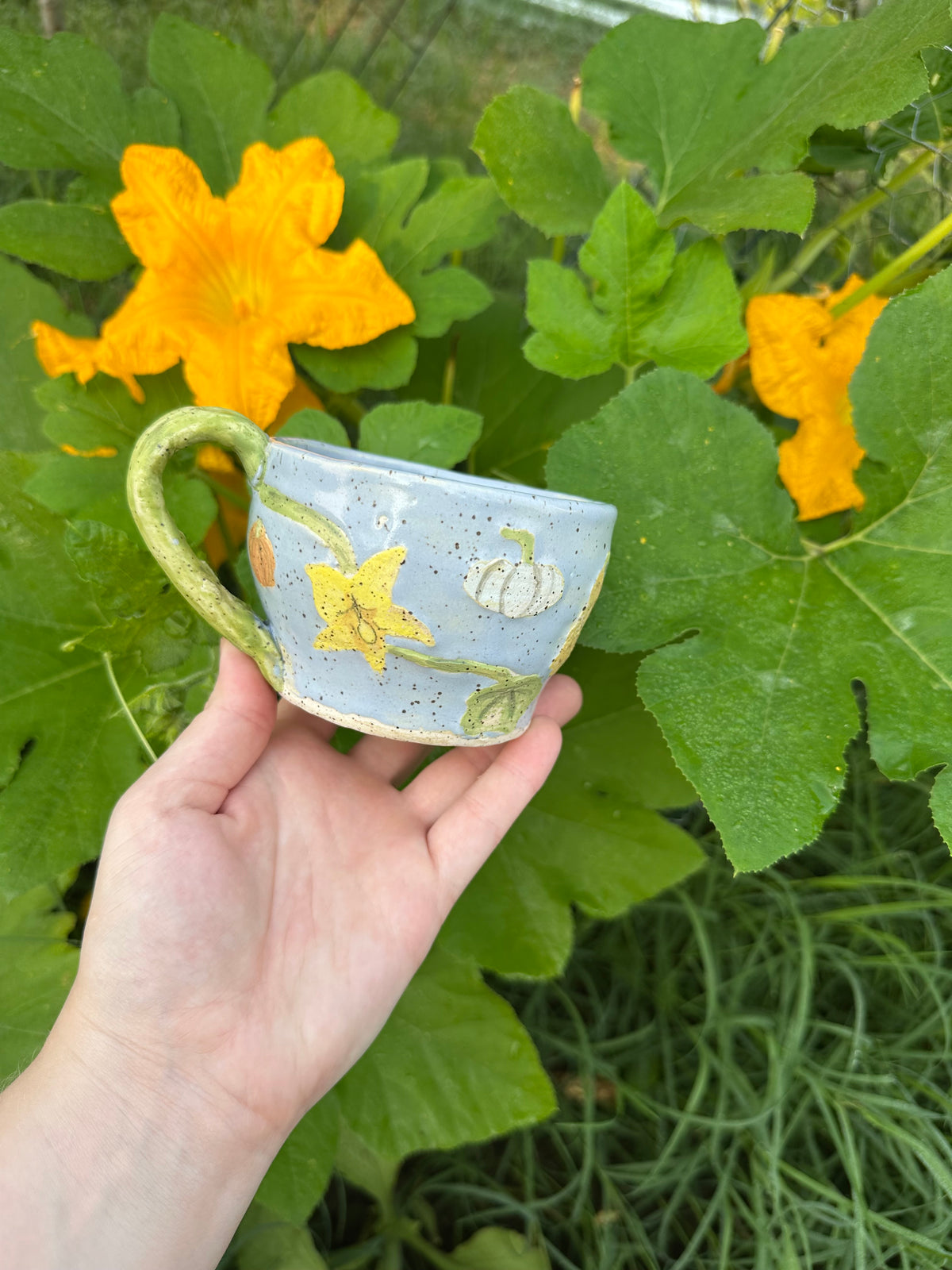 A hand holds a blue ceramic mug with illustrations of pumpkins, pumpkin flowers, pumpkin leaves, and a green handle; set against a garden background of pumpkin leaves and pumpkin flowers.