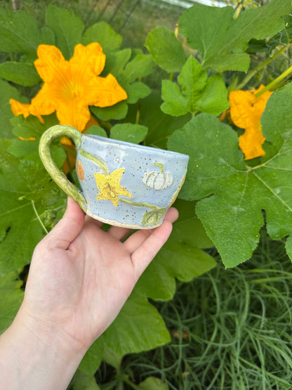 A hand holds a blue ceramic mug with illustrations of pumpkins, pumpkin flowers, pumpkin leaves, and a green handle; set against a garden background of pumpkin leaves and pumpkin flowers.