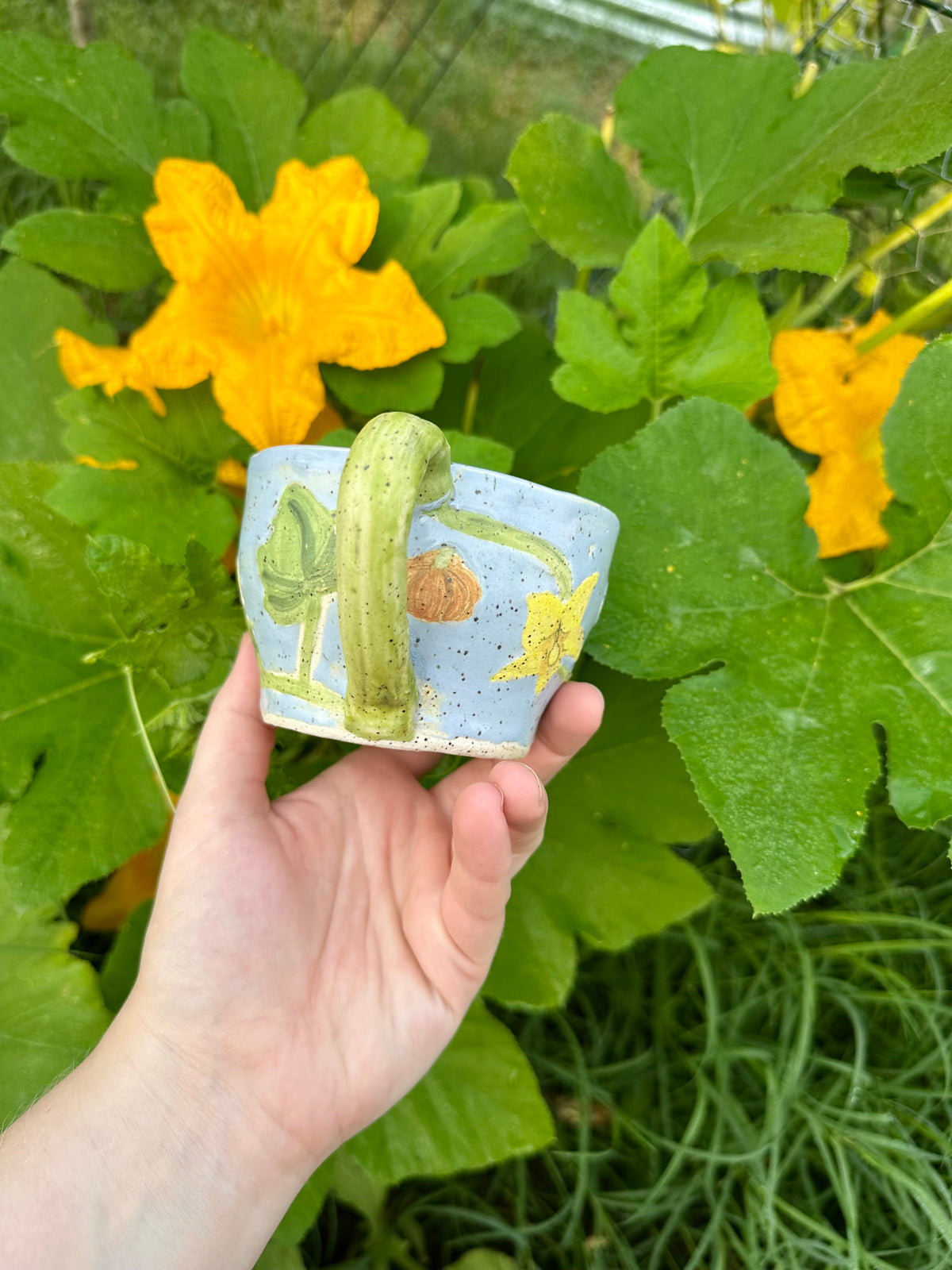 A hand holds a blue ceramic mug with illustrations of pumpkins, pumpkin flowers, pumpkin leaves, and a green handle; set against a garden background of pumpkin leaves and pumpkin flowers.