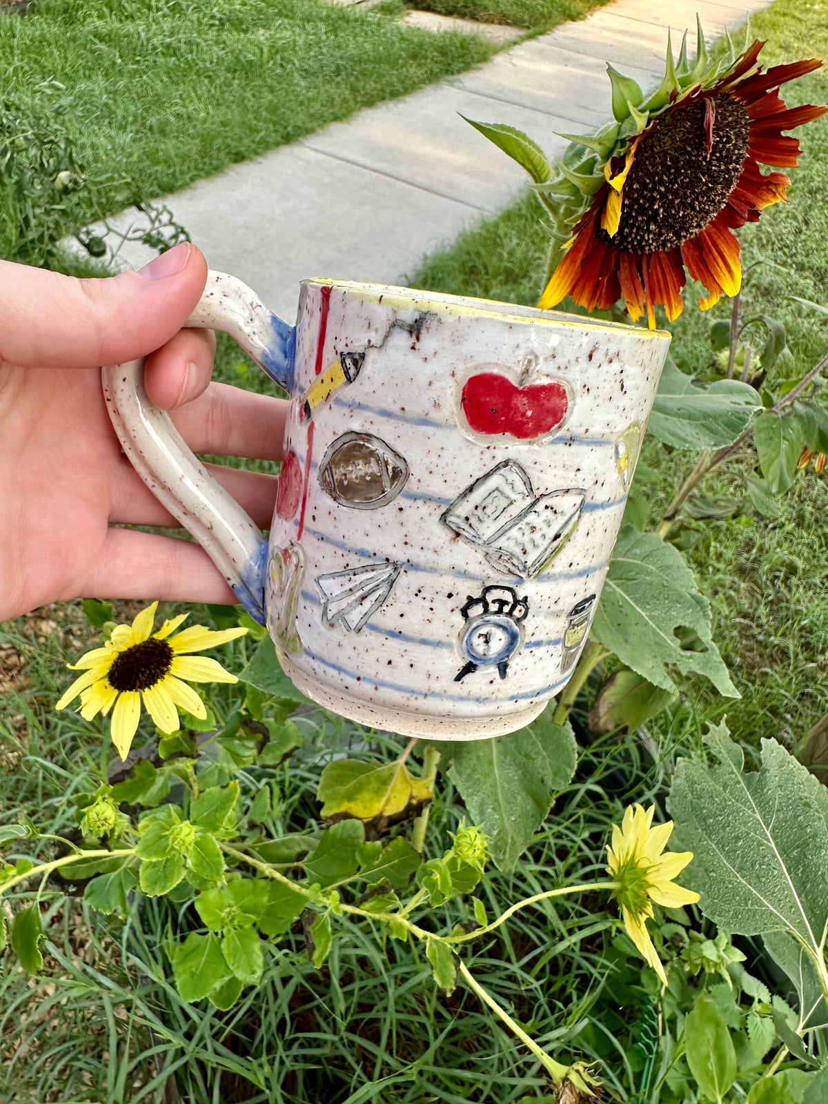 A hand holds a handmade white ceramic mug decorated as a piece of notebook paper with illustrations of books, paper airplanes, apples, pencils, analog alarm clocks, footballs, reading lamps, and to-go coffee cups. Set against a garden background with sunflowers.