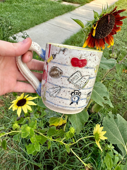 A hand holds a handmade white ceramic mug decorated as a piece of notebook paper with illustrations of books, paper airplanes, apples, pencils, analog alarm clocks, footballs, reading lamps, and to-go coffee cups. Set against a garden background with sunflowers.