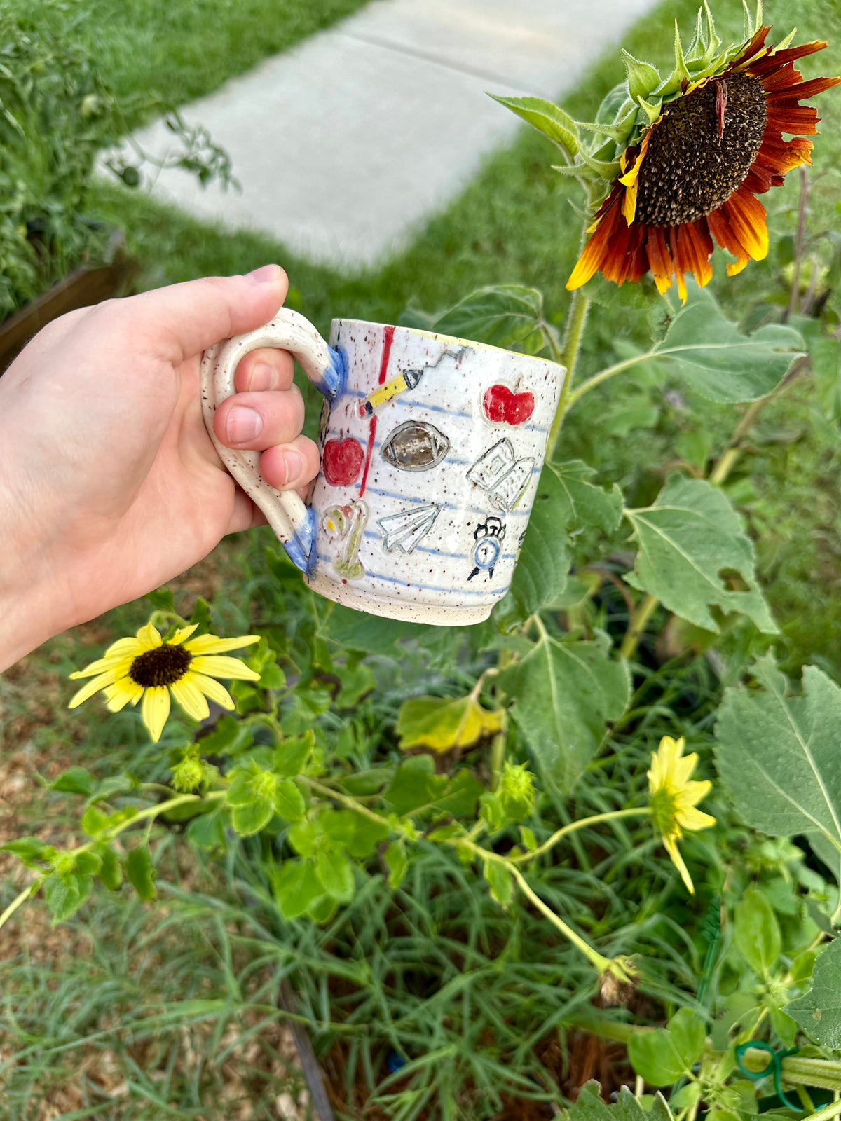 A hand holds a handmade white ceramic mug decorated as a piece of notebook paper with illustrations of books, paper airplanes, apples, pencils, analog alarm clocks, footballs, reading lamps, and to-go coffee cups. Set against a garden background with sunflowers.