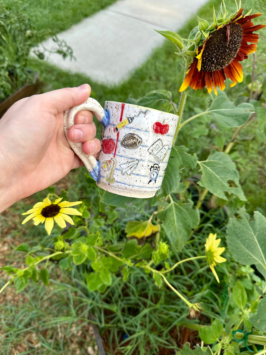 A hand holds a handmade white ceramic mug decorated as a piece of notebook paper with illustrations of books, paper airplanes, apples, pencils, analog alarm clocks, footballs, reading lamps, and to-go coffee cups. Set against a garden background with sunflowers.