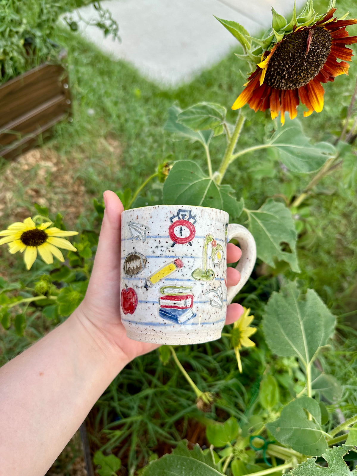 A hand holds a handmade white ceramic mug decorated as a piece of notebook paper with illustrations of books, paper airplanes, apples, pencils, analog alarm clocks, footballs, reading lamps, and to-go coffee cups. Set against a garden background with sunflowers.