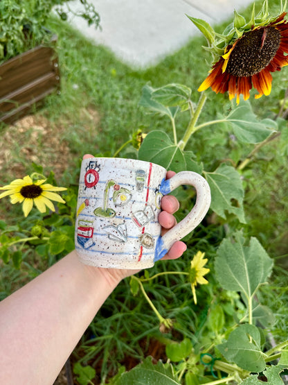 A hand holds a handmade white ceramic mug decorated as a piece of notebook paper with illustrations of books, paper airplanes, apples, pencils, analog alarm clocks, footballs, reading lamps, and to-go coffee cups. Set against a garden background with sunflowers.