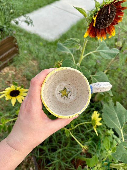 A hand holds a handmade white ceramic mug with yellow rim. Bottom of inside of mug has a happy gold star illustration. Set against a garden background with sunflowers.