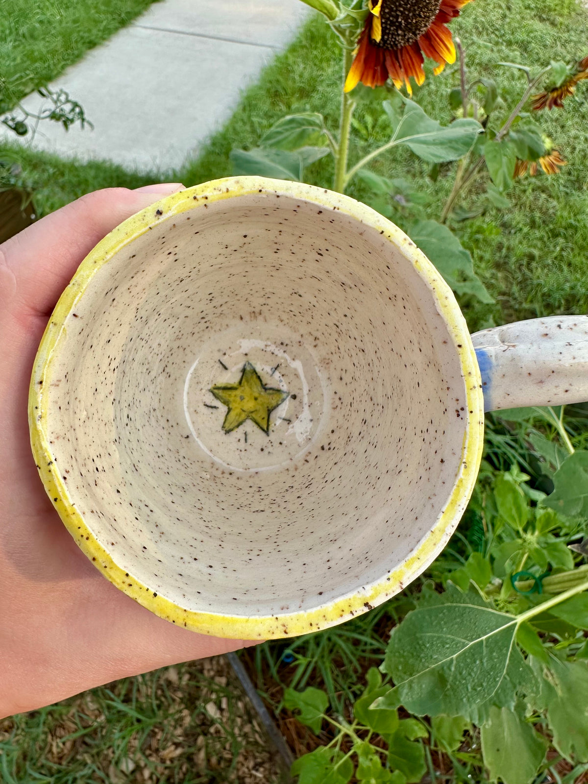 A hand holds a handmade white ceramic mug with yellow rim. Bottom of inside of mug has a happy gold star illustration. Set against a garden background with sunflowers.