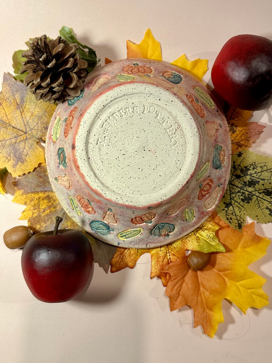 A handmade ceramic bowl with a glossy mauve glaze base, speckled off-white bottom, and a decorated exterior featuring colorful squashes and pumpkins motifs, styled with fall leaves, pinecones, acorns, and red apples on a neutral background.