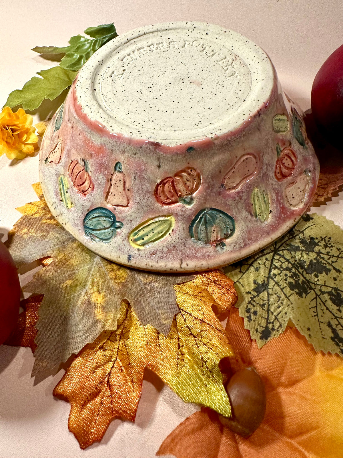 A handmade ceramic bowl with a glossy mauve glaze base, speckled off-white bottom, and a decorated exterior featuring colorful squashes and pumpkins motifs, styled with fall leaves, pinecones, acorns, and red apples on a neutral background.