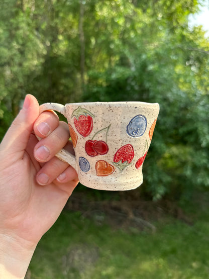 A hand holding a small speckled beige ceramic mug by the handle; the tea cup is decorated with painted red cherries, blueberries, strawberries and peaches, set against a blurred green outdoor background.