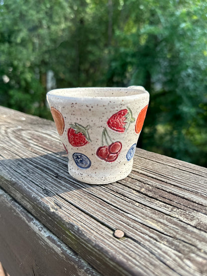A speckled beige ceramic tea cup is decorated with painted red cherries, blueberries, strawberries and peaches, sitting on a worn wood rail set against a blurred green outdoor background.
