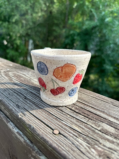 A speckled beige ceramic tea cup is decorated with painted red cherries, blueberries, strawberries and peaches, sitting on a worn wood rail set against a blurred green outdoor background.