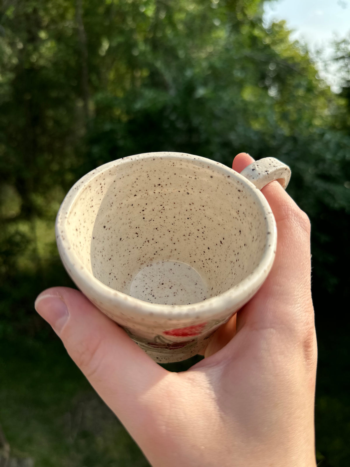 Interior of a speckled beige ceramic mug held in a hand against a blurred green outdoor background.