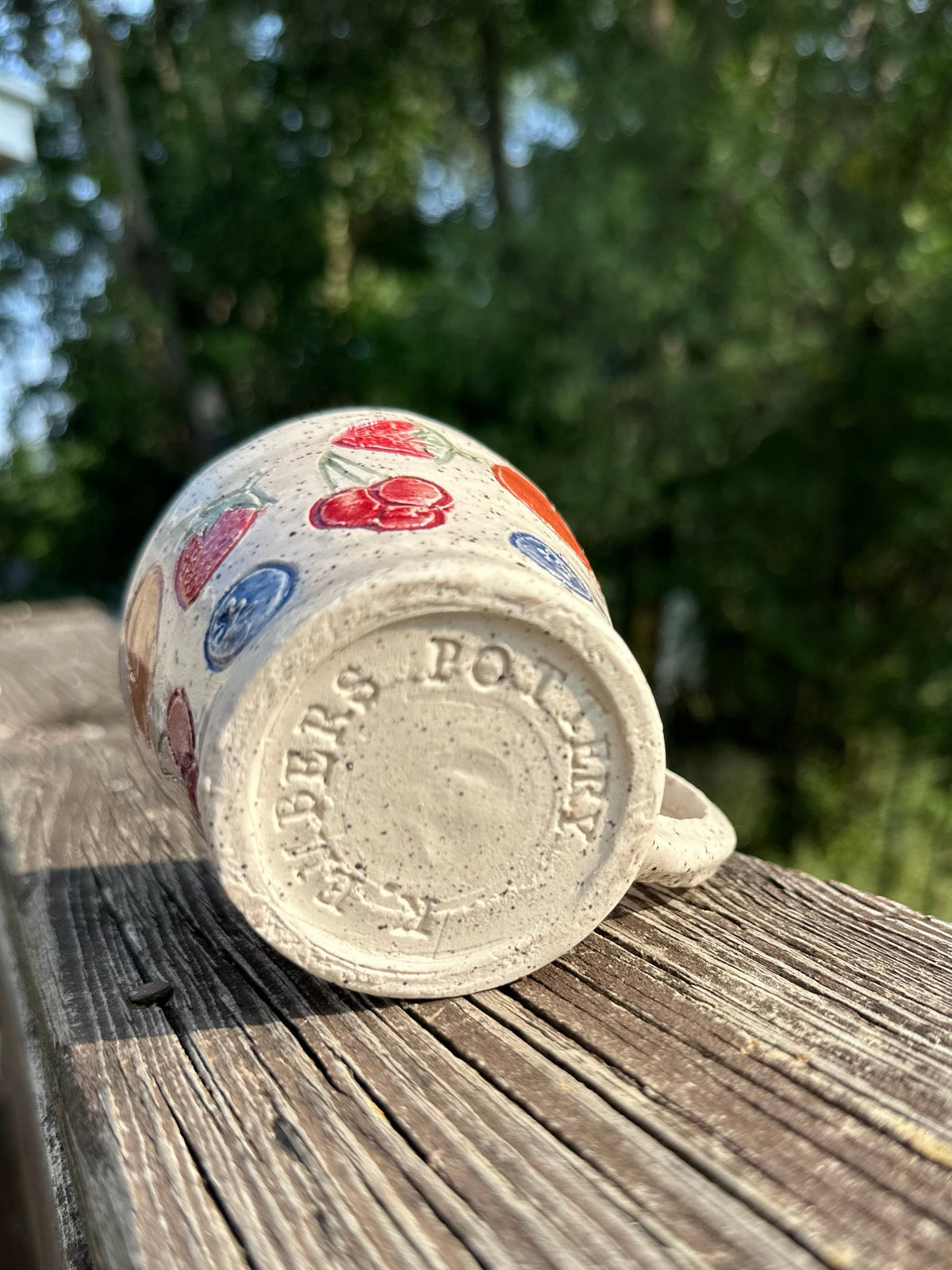 A small speckled beige ceramic mug on its side to show the bottom of the tea cup, stamped with "K. Embers Pottery;" the tea cup is decorated with painted red cherries, blueberries, strawberries and peaches, sitting on a worn wood rail set against a blurred green outdoor background.