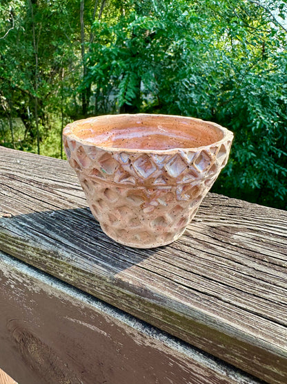 A small-medium bowl carved and colored to look like a waffle cone bowl; slight glossy glaze swirls on the brim and inside makes the illusion of dripping vanilla ice cream. Set on a worn wooden rail in front of a green forest background.